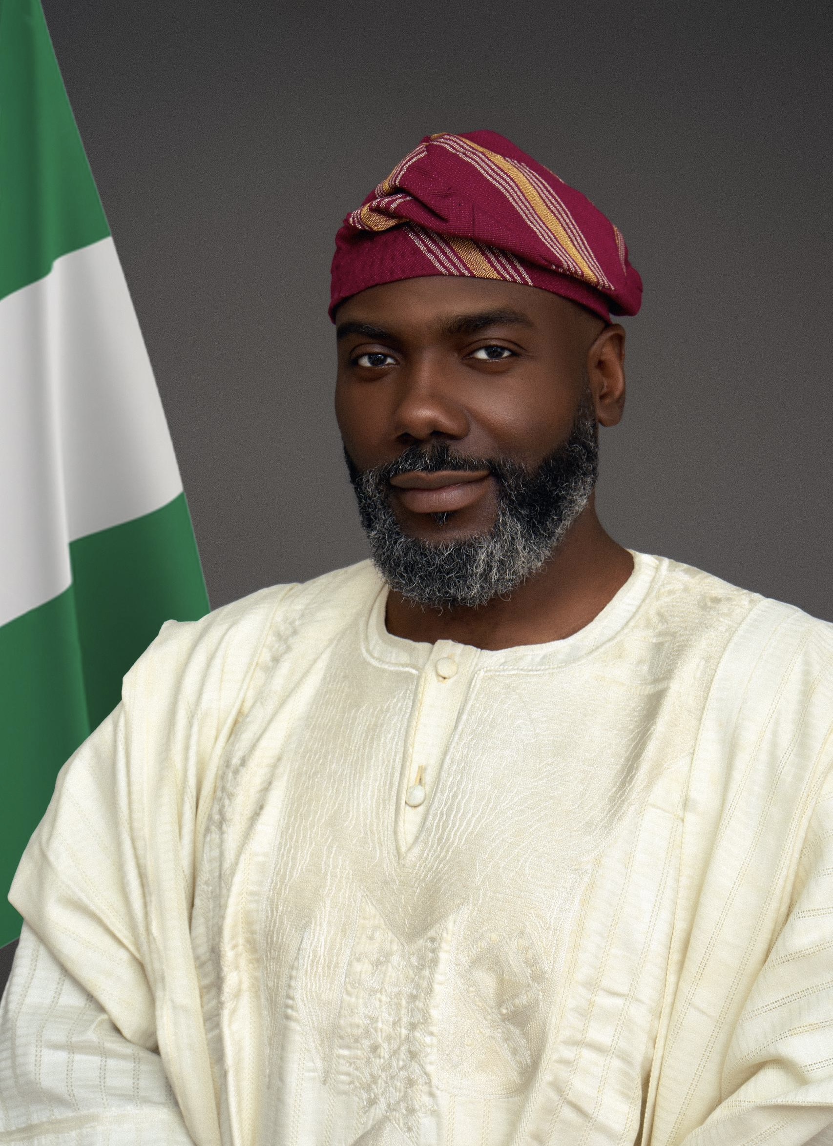 Olugbile Holloway, Director-General of the National Commission for Museums and Monuments (NCMM), wearing traditional white Agbada and a magenta striped Fila cap beside the Nigerian flag
