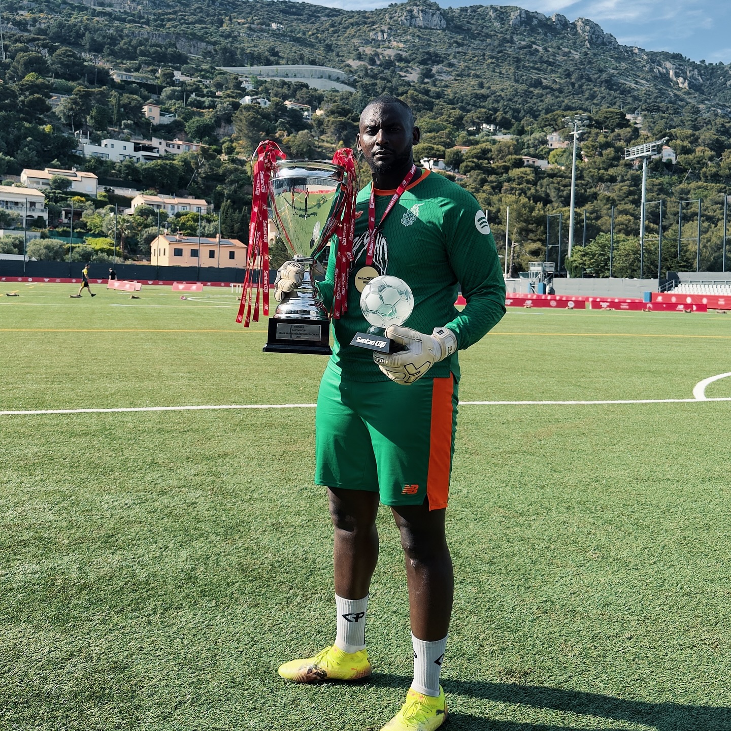 George Desmond Kamurasi in a green goalkeeper kit holding the Santan Cup trophy and match award on a football pitch.
