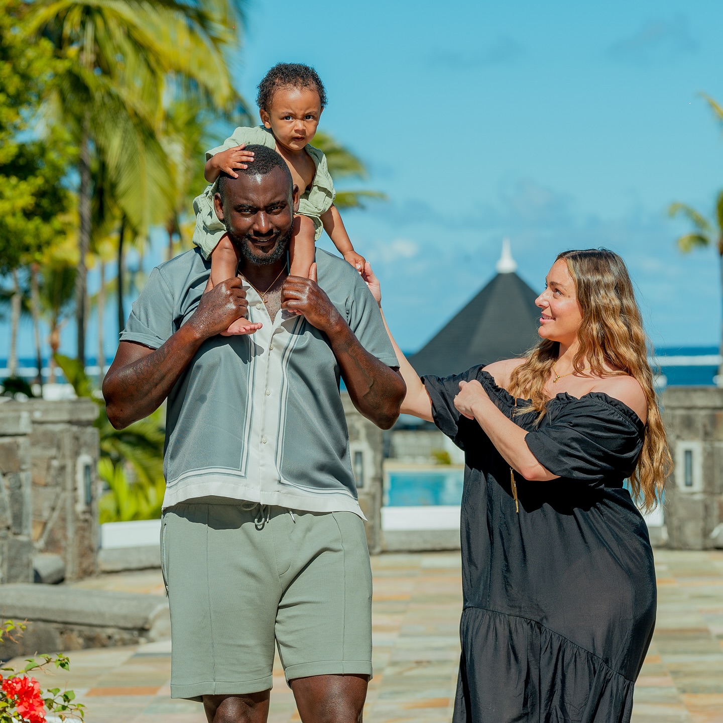 SE Dons captain George Desmond Kamurasi holding his young son on his shoulders alongside his wife Abbey Kamurasi outdoors.