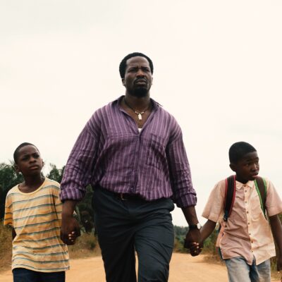 British-Nigerian actor Ṣọpẹ́ Dìrísù as Folarin holding hands with young sons Remi and Akin on a dirt road in My Father’s Shadow.