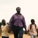 British-Nigerian actor Ṣọpẹ́ Dìrísù as Folarin holding hands with young sons Remi and Akin on a dirt road in My Father’s Shadow.