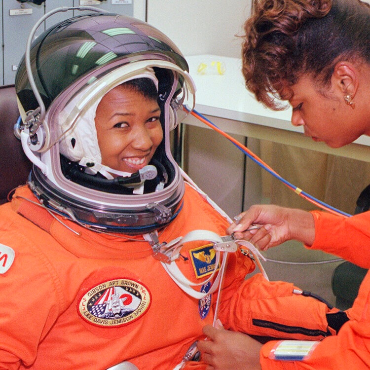 Technician assisting Dr Mae Jemison with her orange flight suit helmet harness.