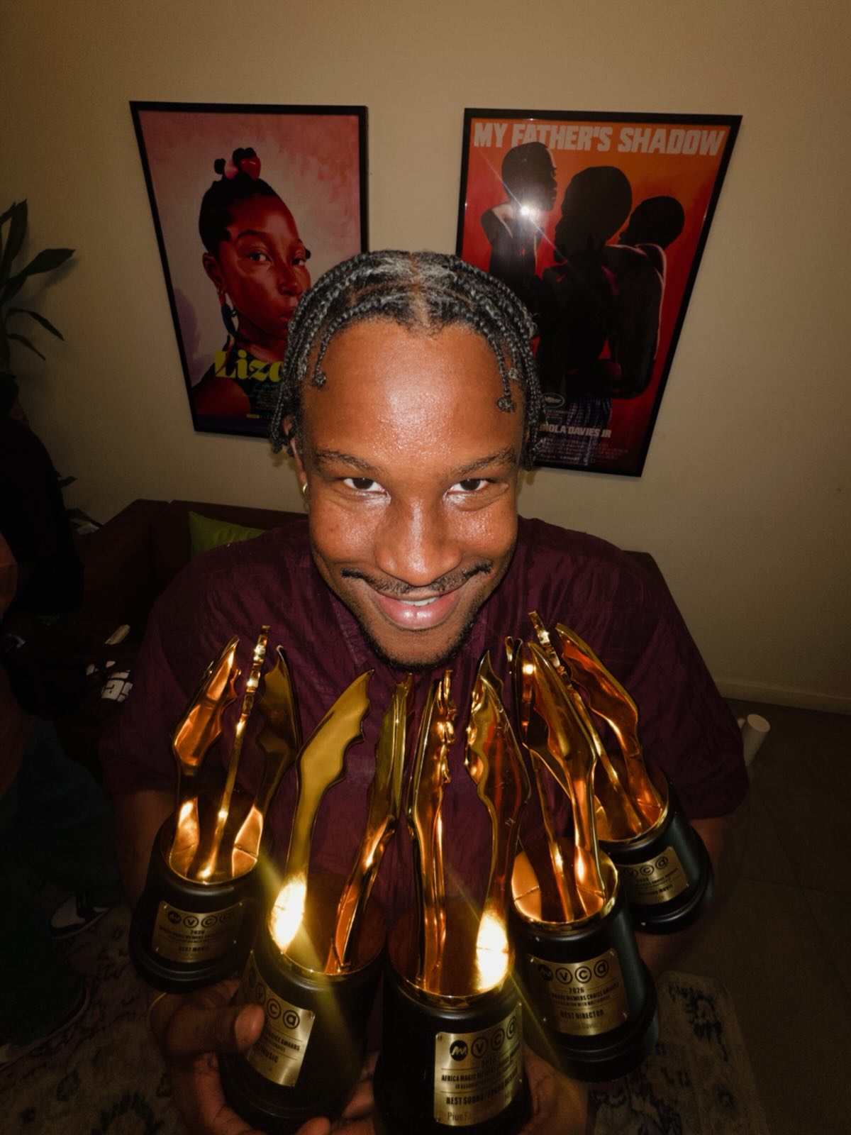 Director Akinola Davies Jr. smiling while holding multiple AMVCA golden trophies with My Father's Shadow poster in background.