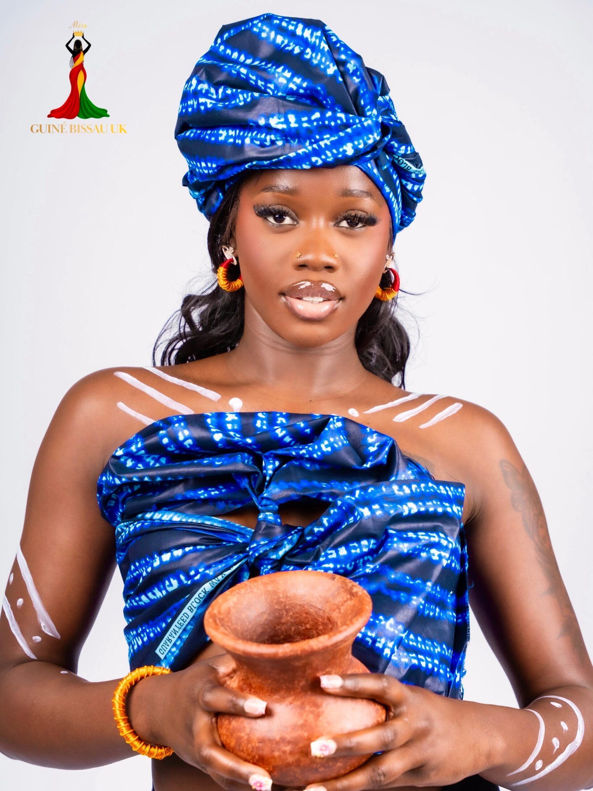 Luiza Santos holding a clay pot while wearing a bright blue printed headwrap and matching top for Miss Guiné-Bissau UK 2026