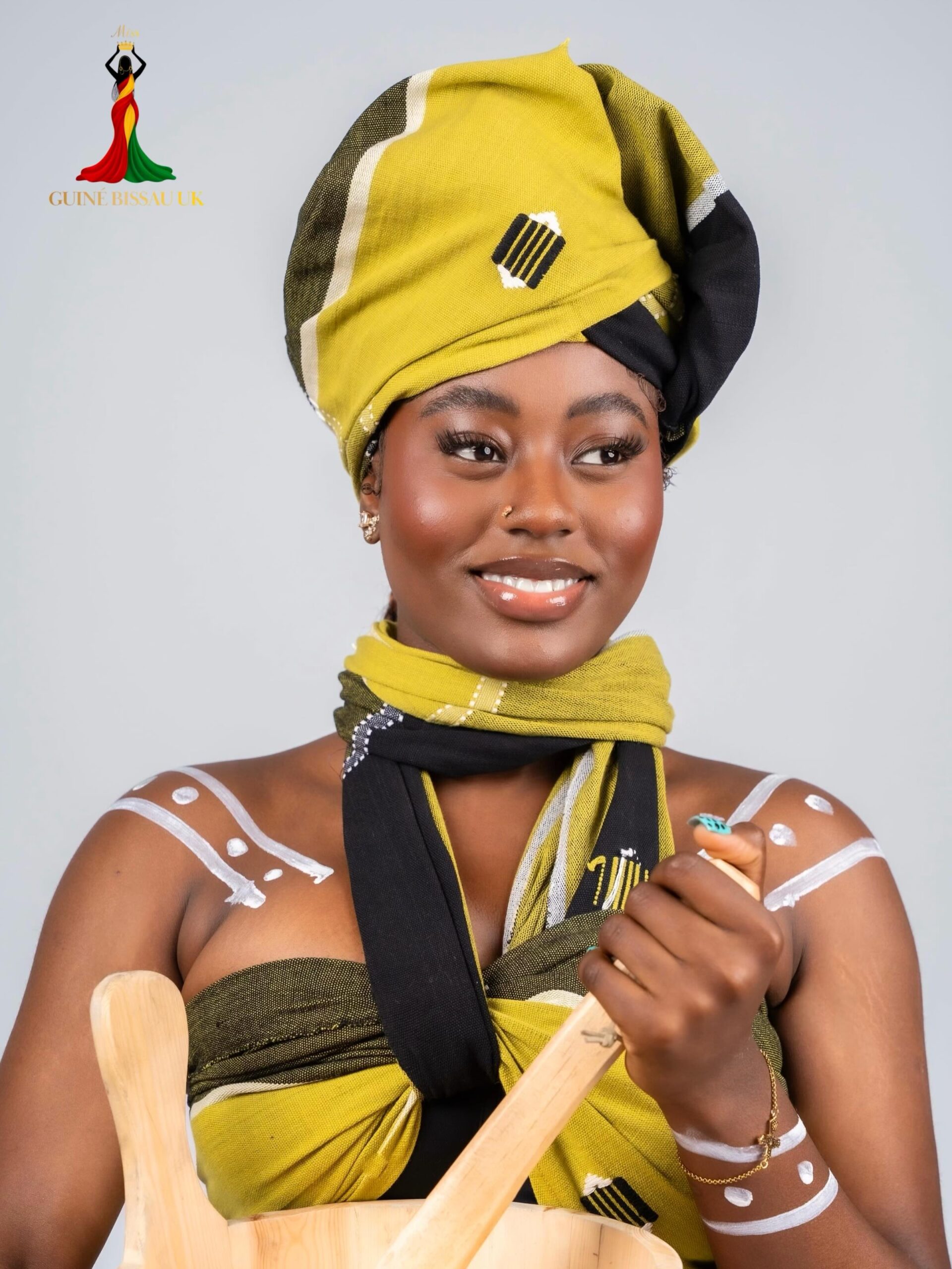 Eldinha Lopes wearing a olive green and black woven headwrap and matching neck scarf holding a wooden mortar for Miss Guiné-Bissau UK 2026