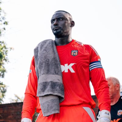 SE Dons captain George Desmond Kamurasi on the touchline wearing a red goalkeeper jersey and captain's armband.