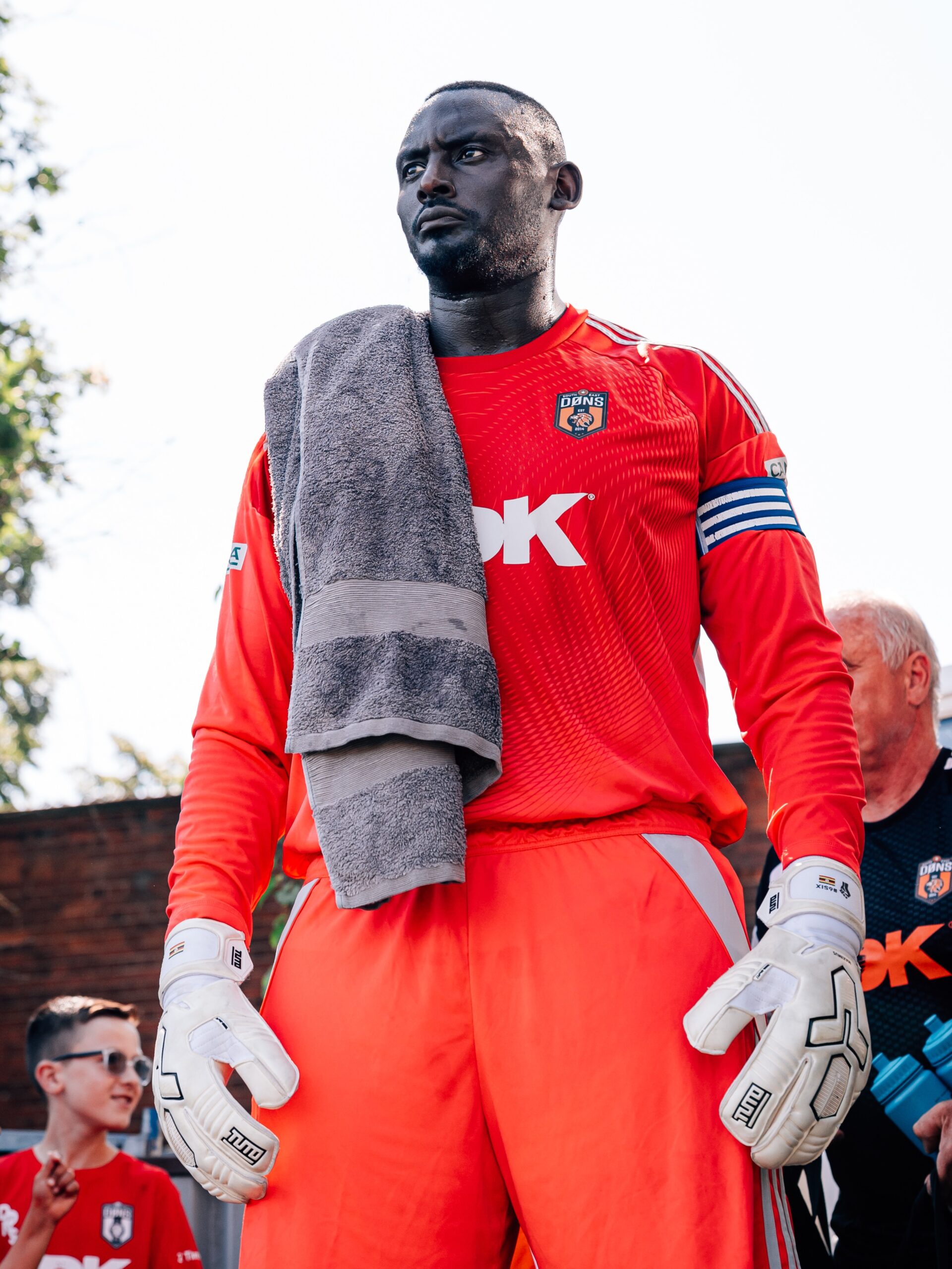 SE Dons captain George Desmond Kamurasi on the touchline wearing a red goalkeeper jersey and captain's armband.