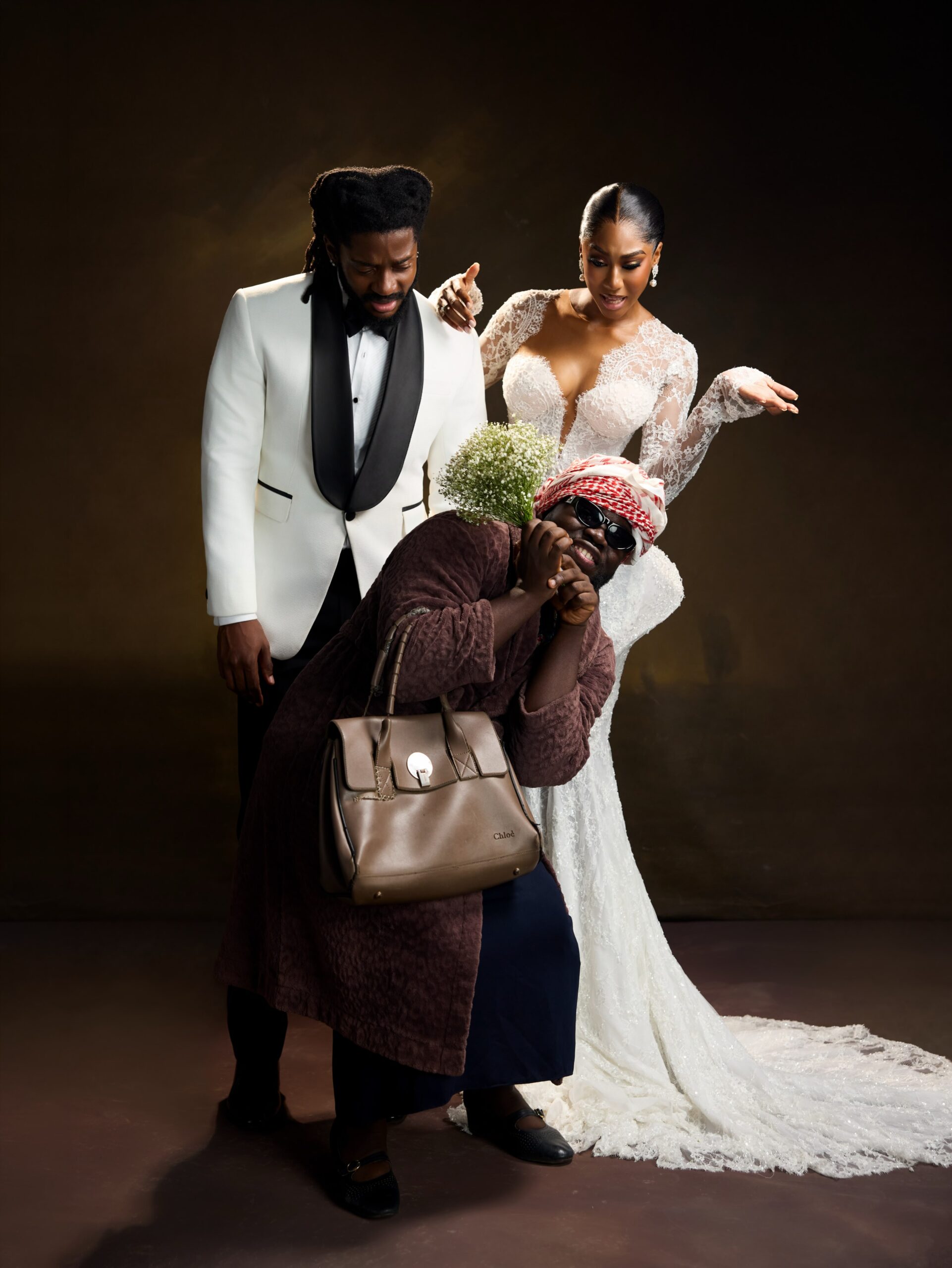 Laura Ajayi in a lace wedding gown and David Uruka in a white tuxedo standing behind Folagade Banks as Mama Deola