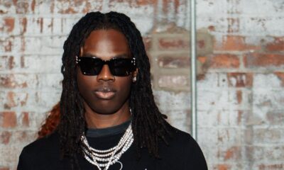 Afrobeats artist Rema wearing black sunglasses, a black long-sleeve shirt with Dreams embroidered, and silver chains.