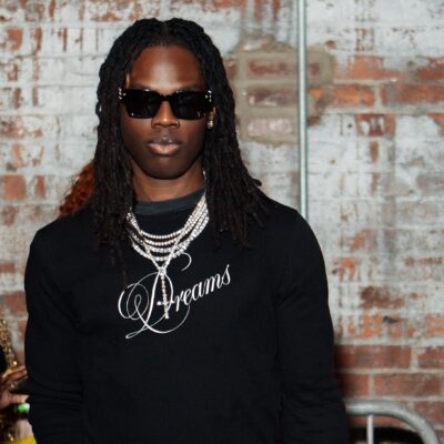 Afrobeats artist Rema wearing black sunglasses, a black long-sleeve shirt with Dreams embroidered, and silver chains.