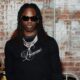 Afrobeats artist Rema wearing black sunglasses, a black long-sleeve shirt with Dreams embroidered, and silver chains.