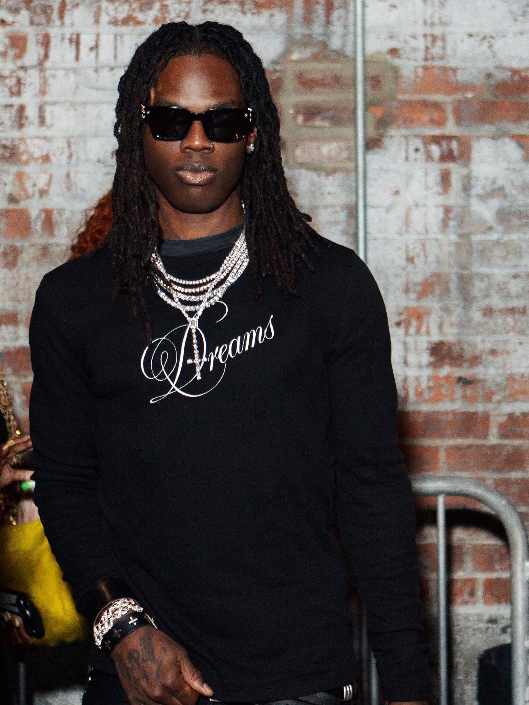 Afrobeats artist Rema wearing black sunglasses, a black long-sleeve shirt with Dreams embroidered, and silver chains.