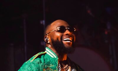 Davido smiling and performing with a microphone on stage in a green lace bomber jacket and sunglasses.