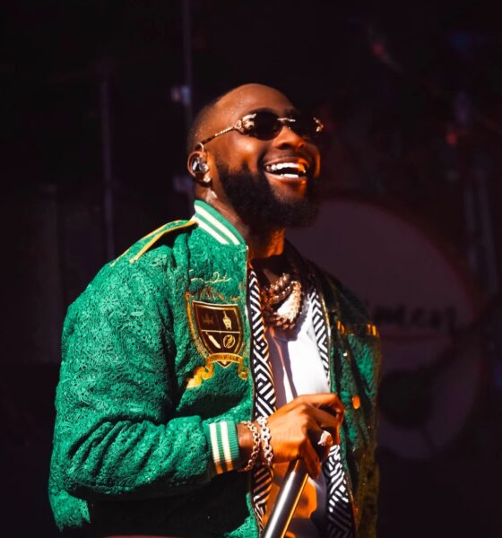 Davido smiling and performing with a microphone on stage in a green lace bomber jacket and sunglasses.