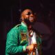 Davido smiling and performing with a microphone on stage in a green lace bomber jacket and sunglasses.