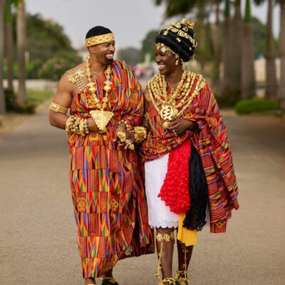 Bozoma Saint John and Keely Watson smiling at each other while walking in traditional hand-woven Kente drapes and gold jewellery.