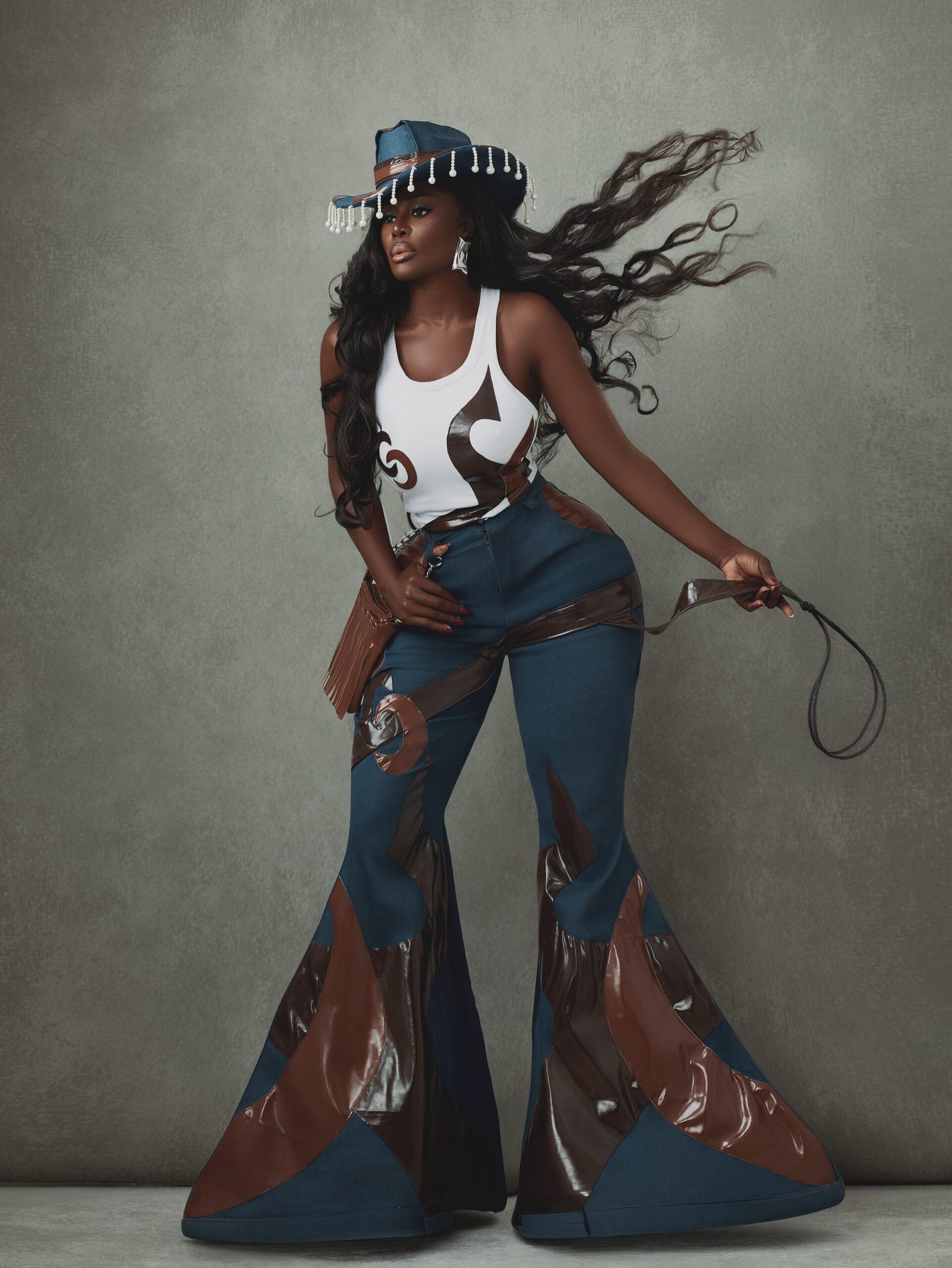 Ghanaian fashion icon Nana Akua Addo posing in flared denim trousers with brown leather flame patches, a white ribbed tank top, metallic-fringed cowboy hat, and leather whip.