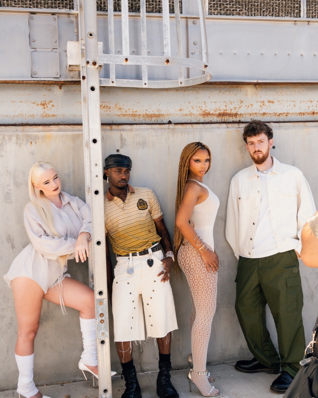 Clean Bandit, Omah Lay, and Chlöe Bailey standing against a grey metal wall beside a vertical safety ladder.