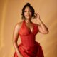 Joselyn Dumas posing with a high top-knot updo hairstyle and glossy nude lip makeup wearing a red halterneck corset dress