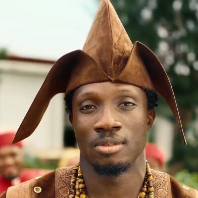 Afrogospel artist Greatman Takit wearing a brown Yoruba agbada, beaded neckpiece, and traditional fila cap on the set of his ADML music video.
