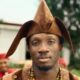 Afrogospel artist Greatman Takit wearing a brown Yoruba agbada, beaded neckpiece, and traditional fila cap on the set of his ADML music video.