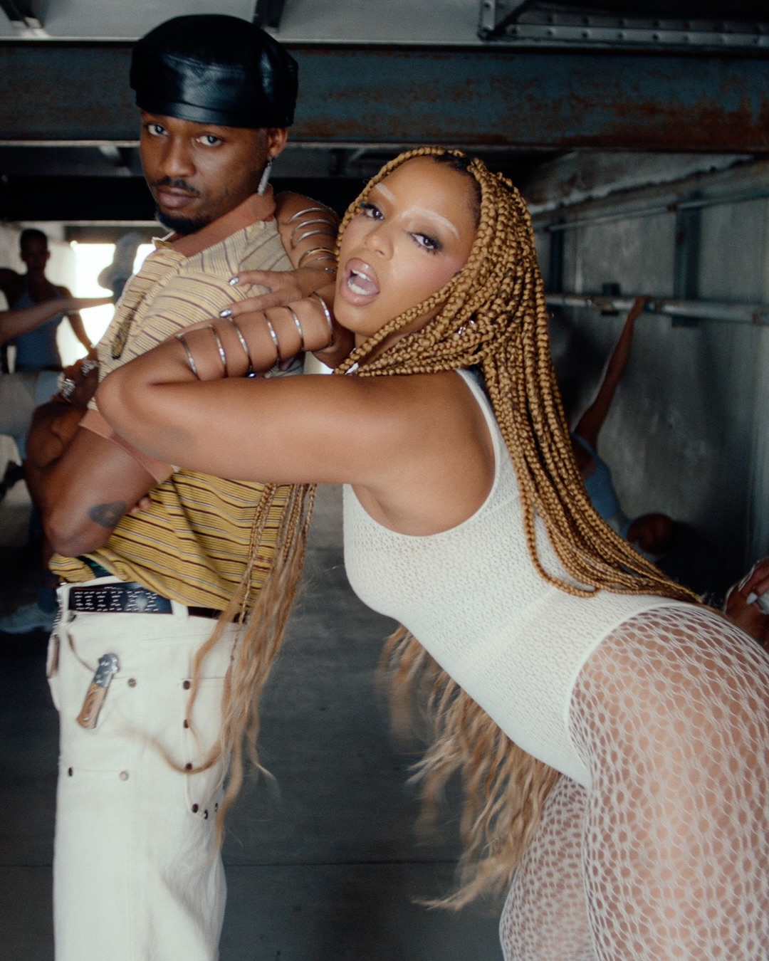 Chlöe Bailey leaning her head on Omah Lay's shoulder while he stands in a striped polo shirt, leather cap, and wide-leg trousers.