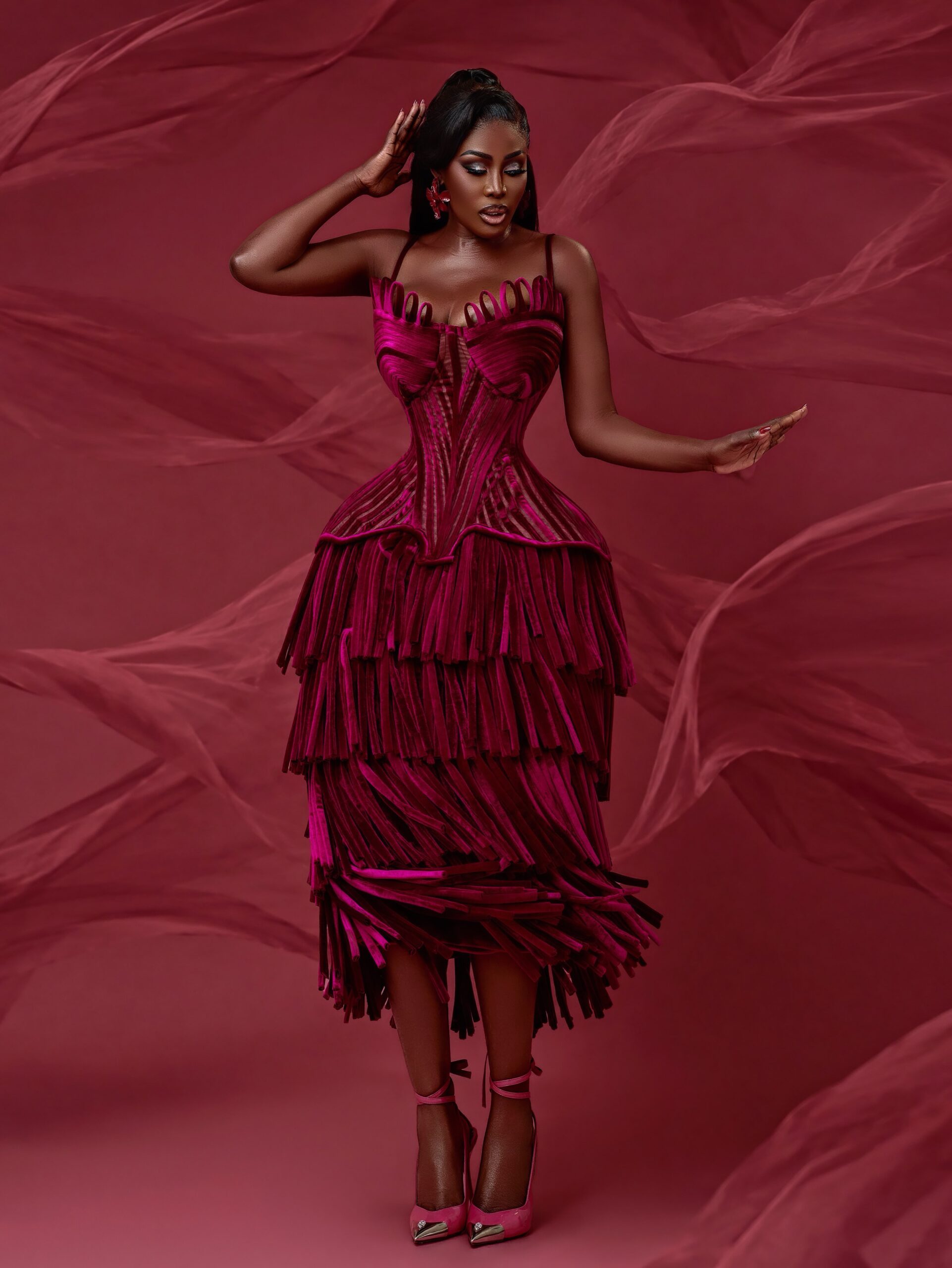 Nana Akua Addo posing in a rich magenta-crimson midi dress with a corseted ribbed bodice and three tiered layers of fine velvet fringe.
