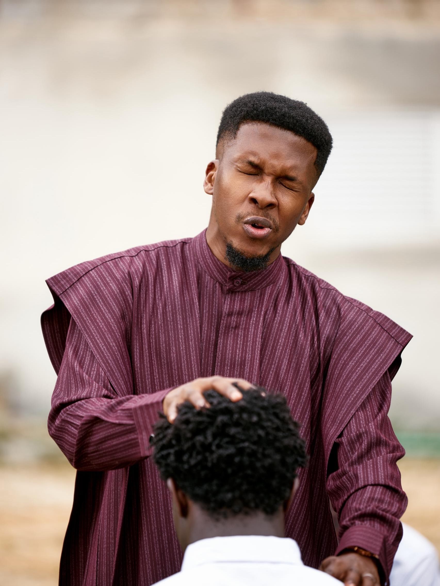 Pastor Emmanuel Iren praying and laying hands on Greatman Takit's head during a scene in the ADML music video.