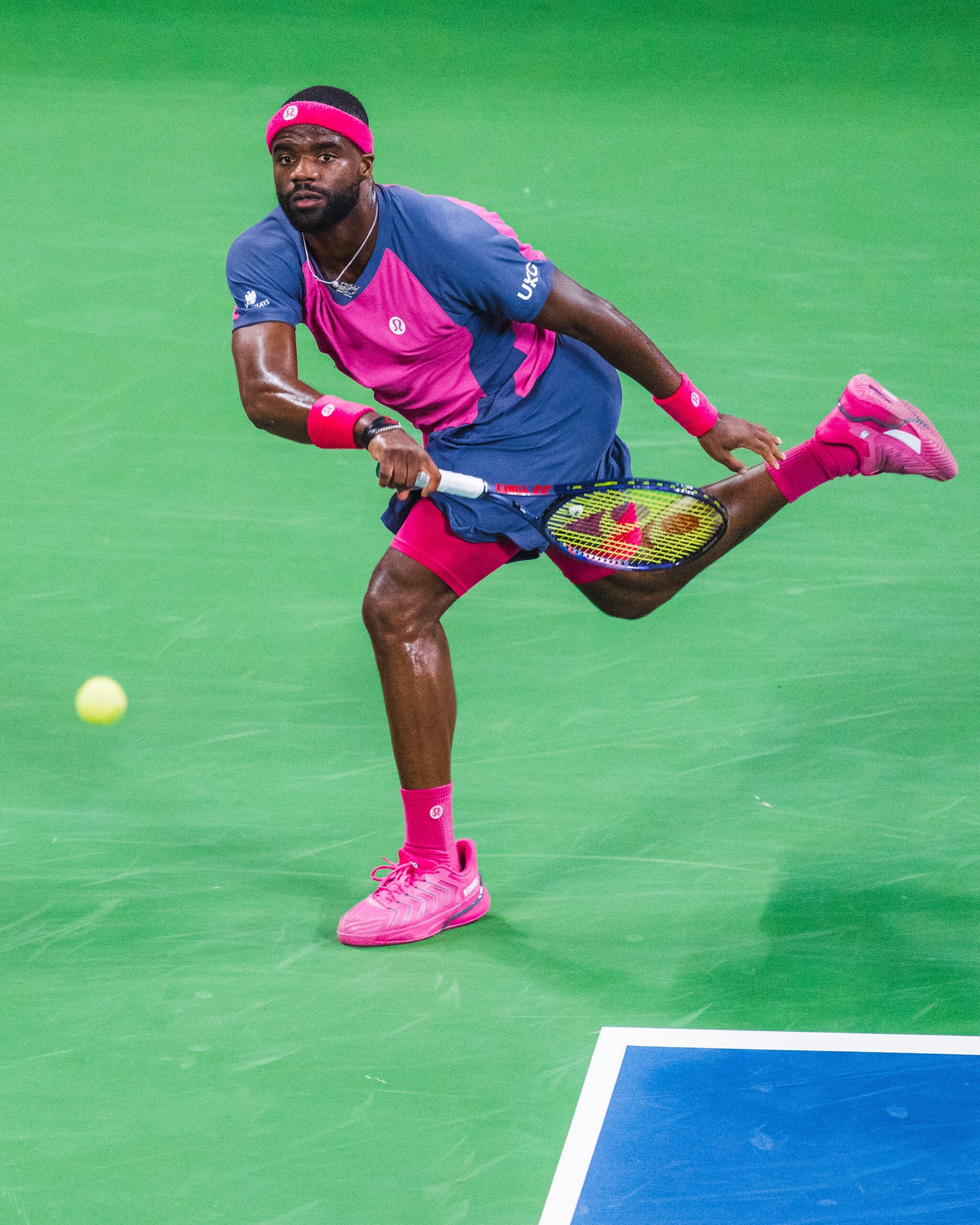 Frances Tiafoe running to strike a forehand return in a pink and navy Lululemon kit.