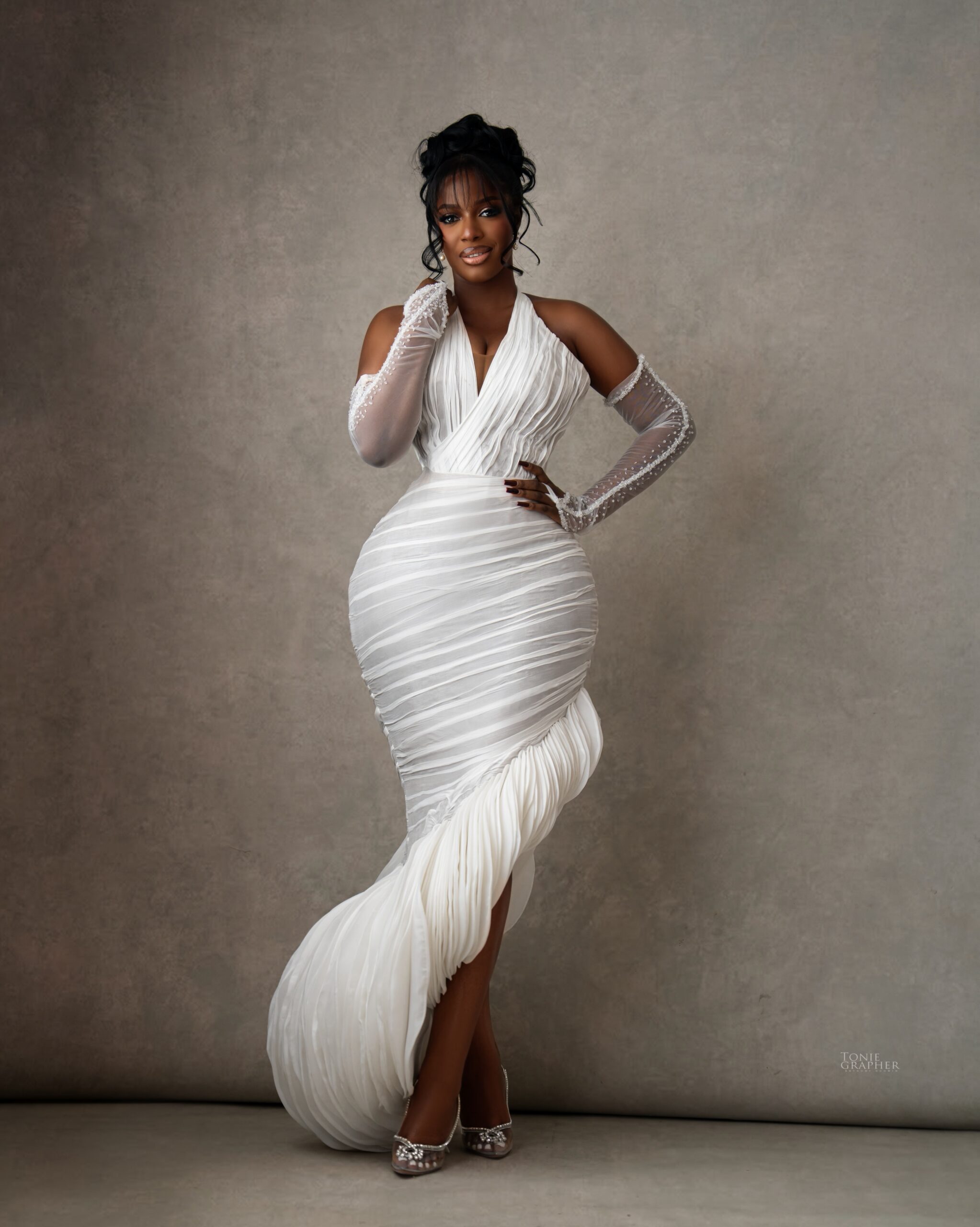 Hilda Baci posing in a white ruched halterneck mermaid dress with sheer pearl opera gloves at A Mama Deola Wedding Story premiere.