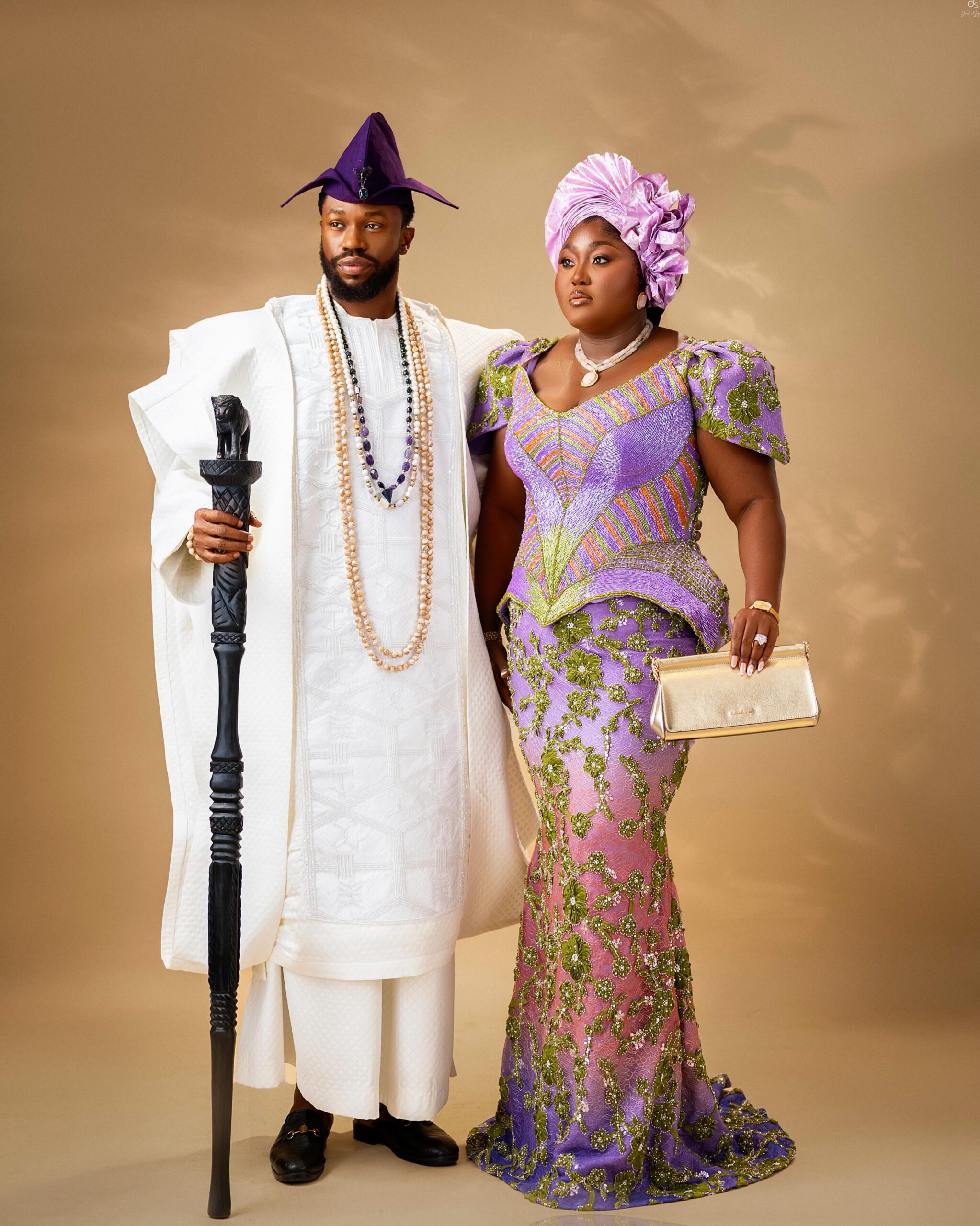 Stan Nze in a white embroidered Agbada holding a lion-head staff beside Blessing Nze in an ombré purple gown and lilac Gele.
