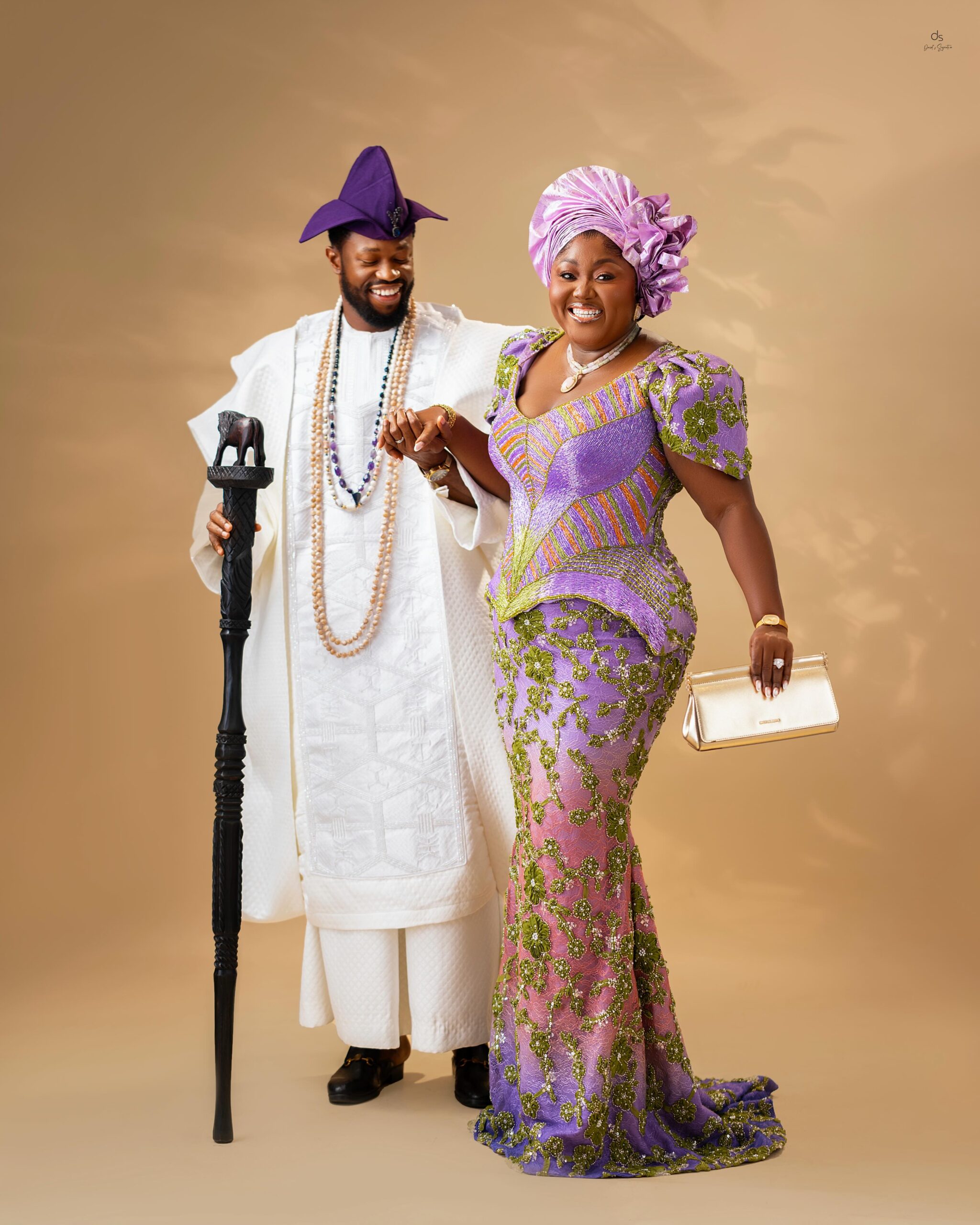 Stan Nze holding hands with Blessing Nze as they smile together in matching white and purple traditional Nigerian outfits.