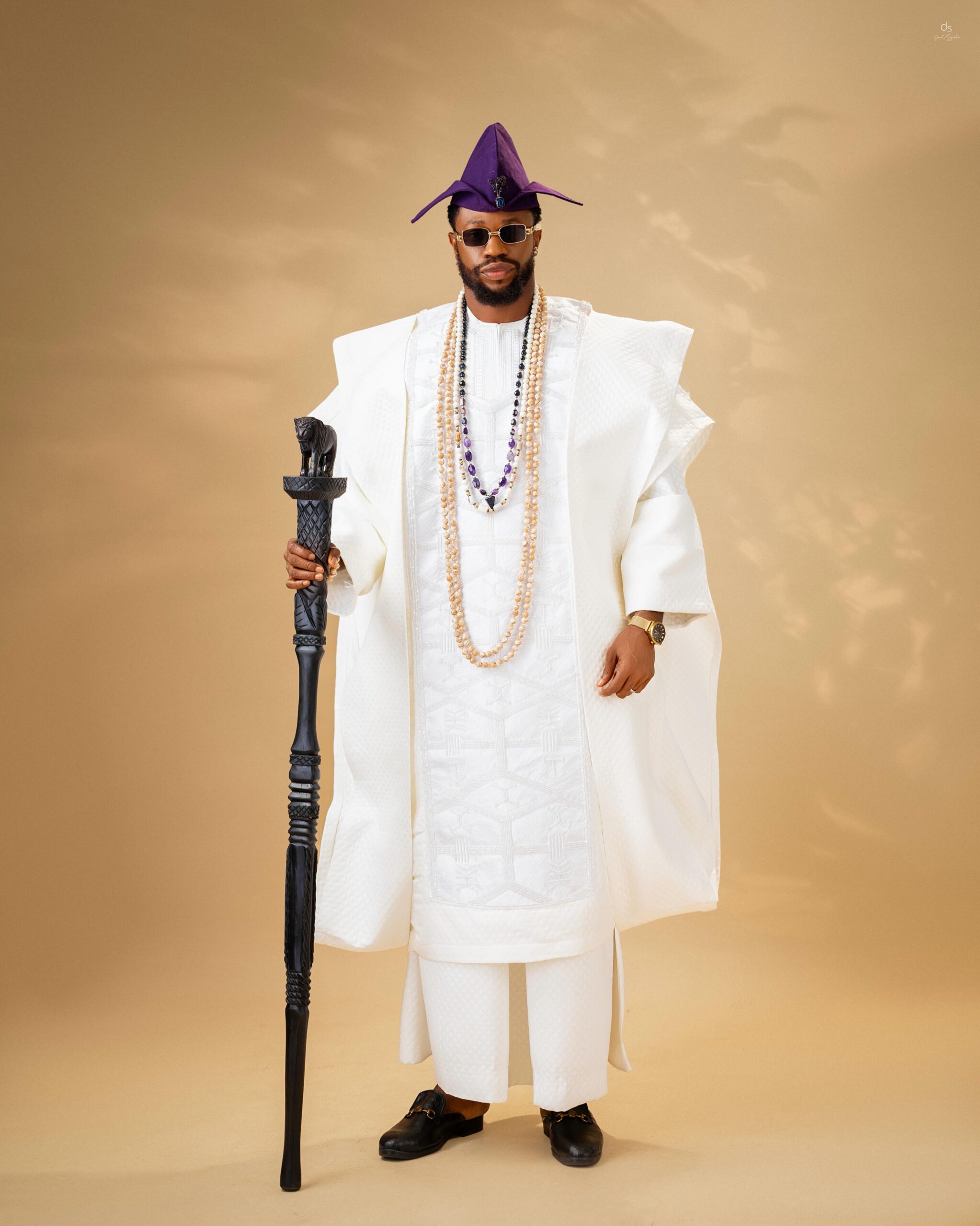 Stan Nze posing in a white embroidered Agbada set with a deep purple Fila cap, dark sunglasses, and a carved black lion-head wooden staff.