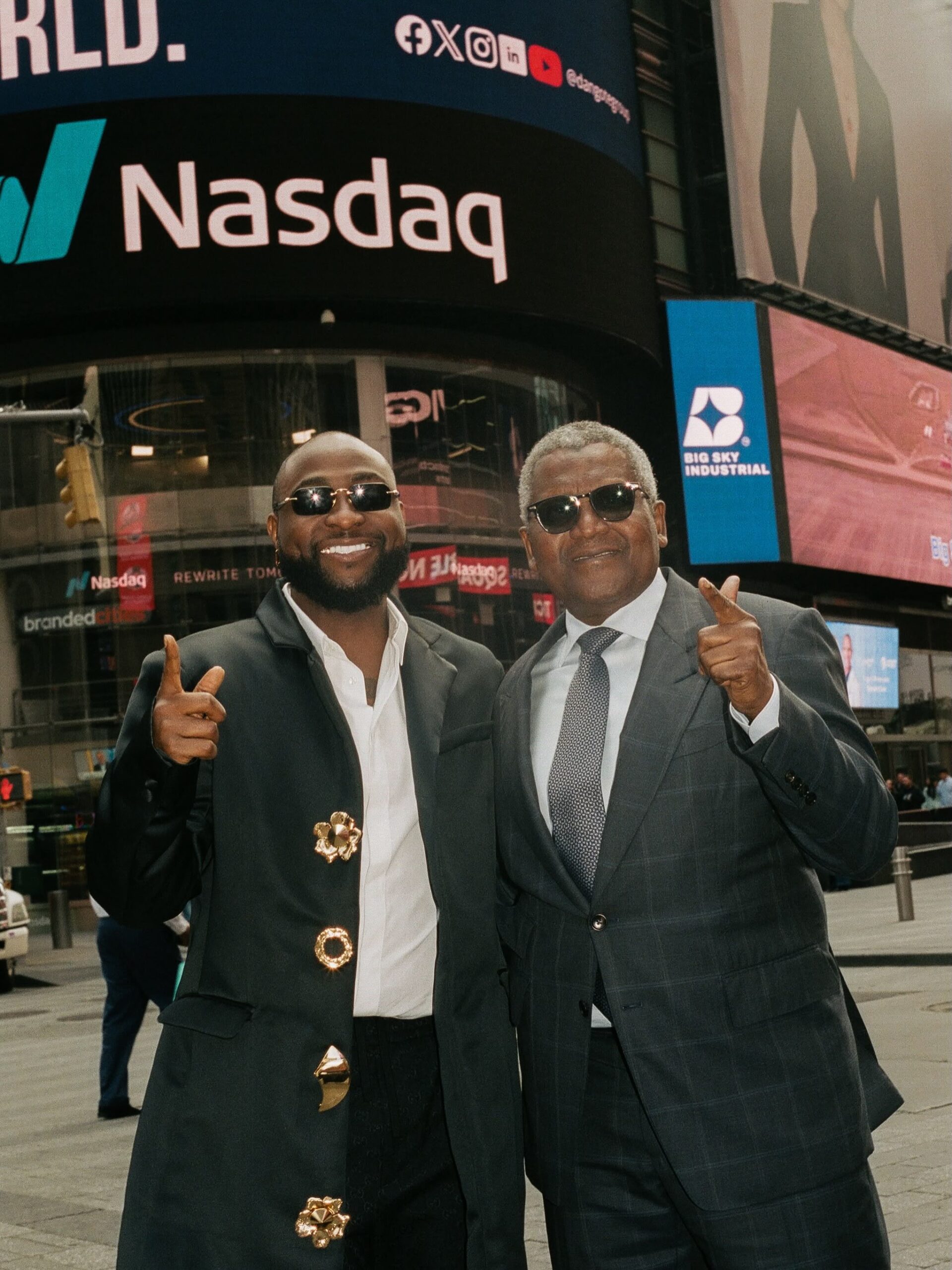 Davido and Aliko Dangote posing with thumbs-up gestures beneath the Nasdaq billboard in Times Square New York