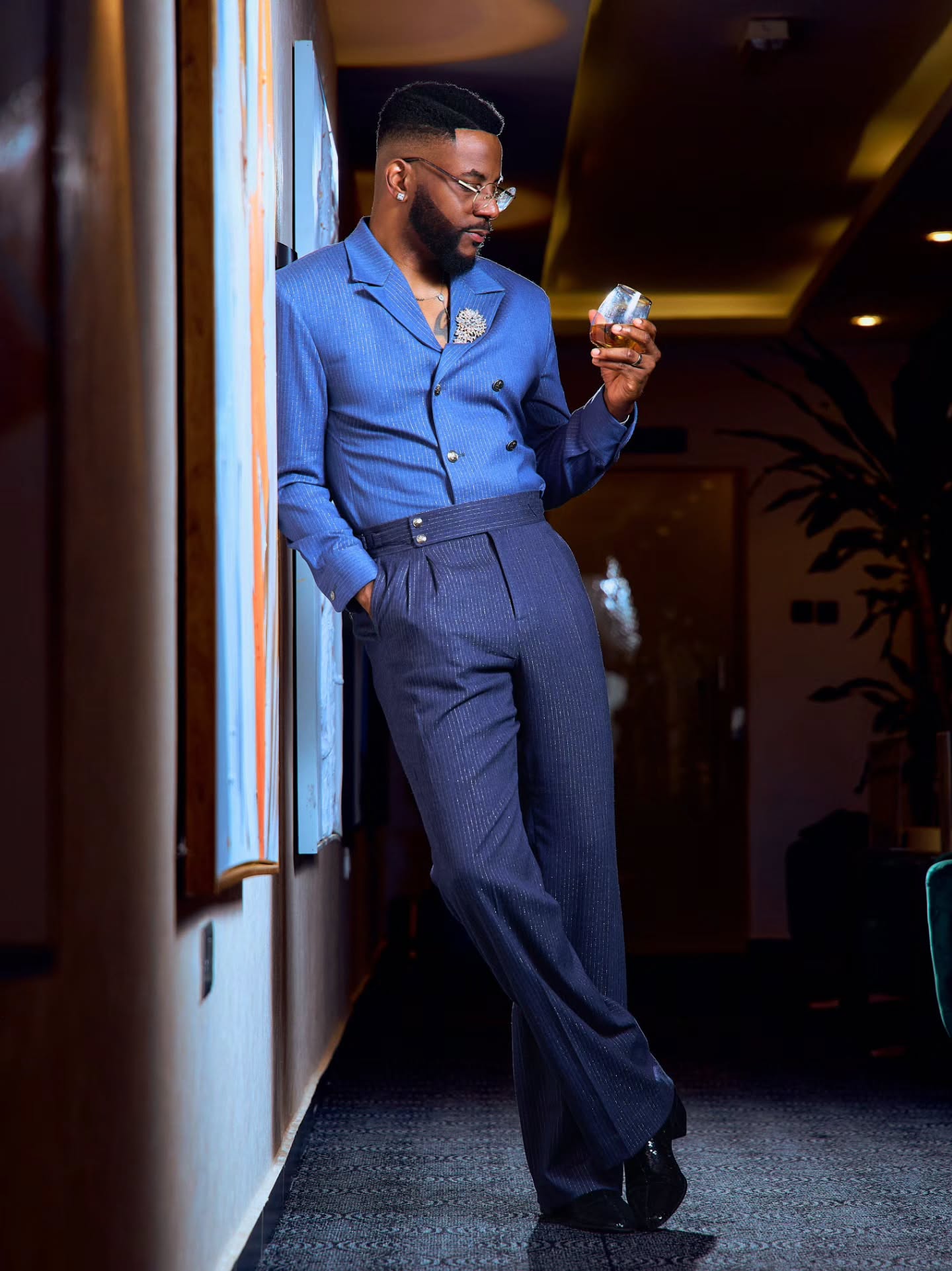 Ebuka Obi-Uchendu holding a glass in a custom blue double-breasted crossover shirt-jacket and high-waisted navy Gurkha trousers by Atafo.