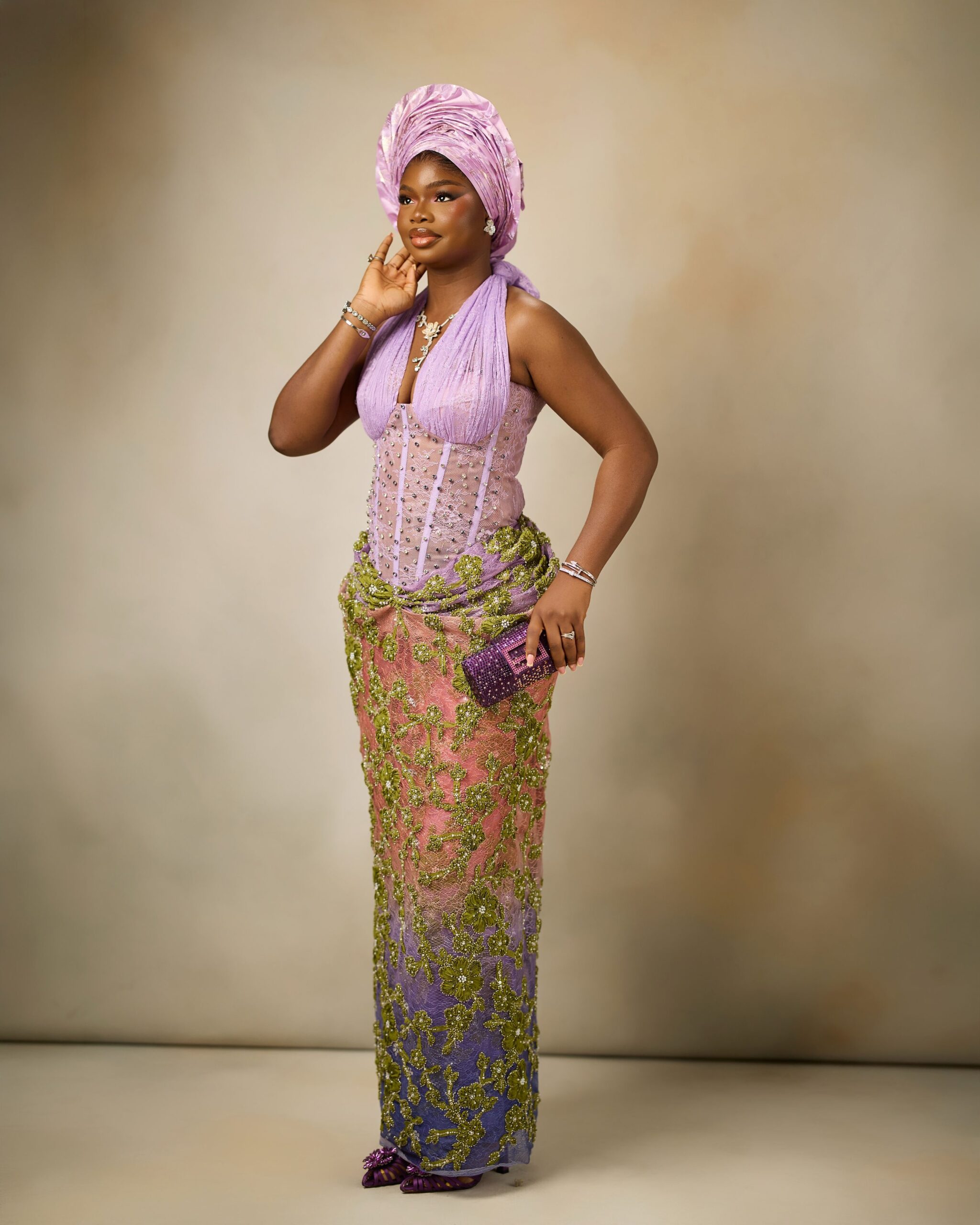 Olamide posing in a lilac corset halter dress with an olive green floral lace skirt and metallic gele.