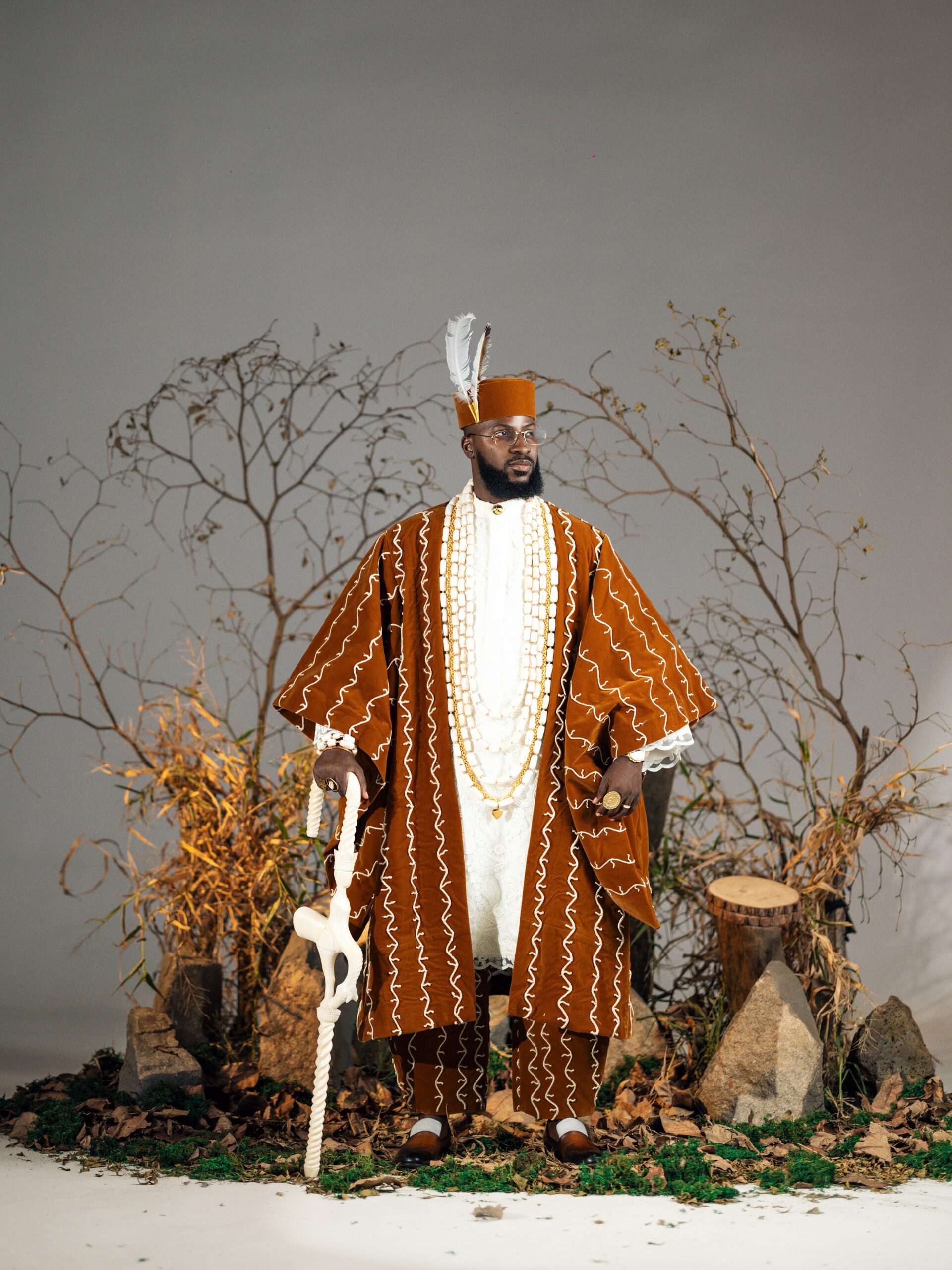 Akin Faminu wearing a brown velvet embroidered Agbada by Deji and Kola with coral beads and staff.