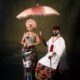Kiky Festus under a pink parasol next to Akin Faminu in a white tunic and tartan George skirt.