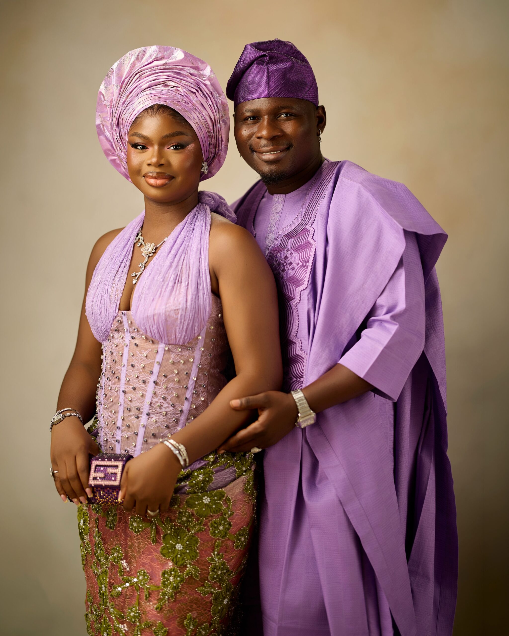 Olamide holding a purple box clutch standing beside Kamo State in an embroidered lilac agbada.