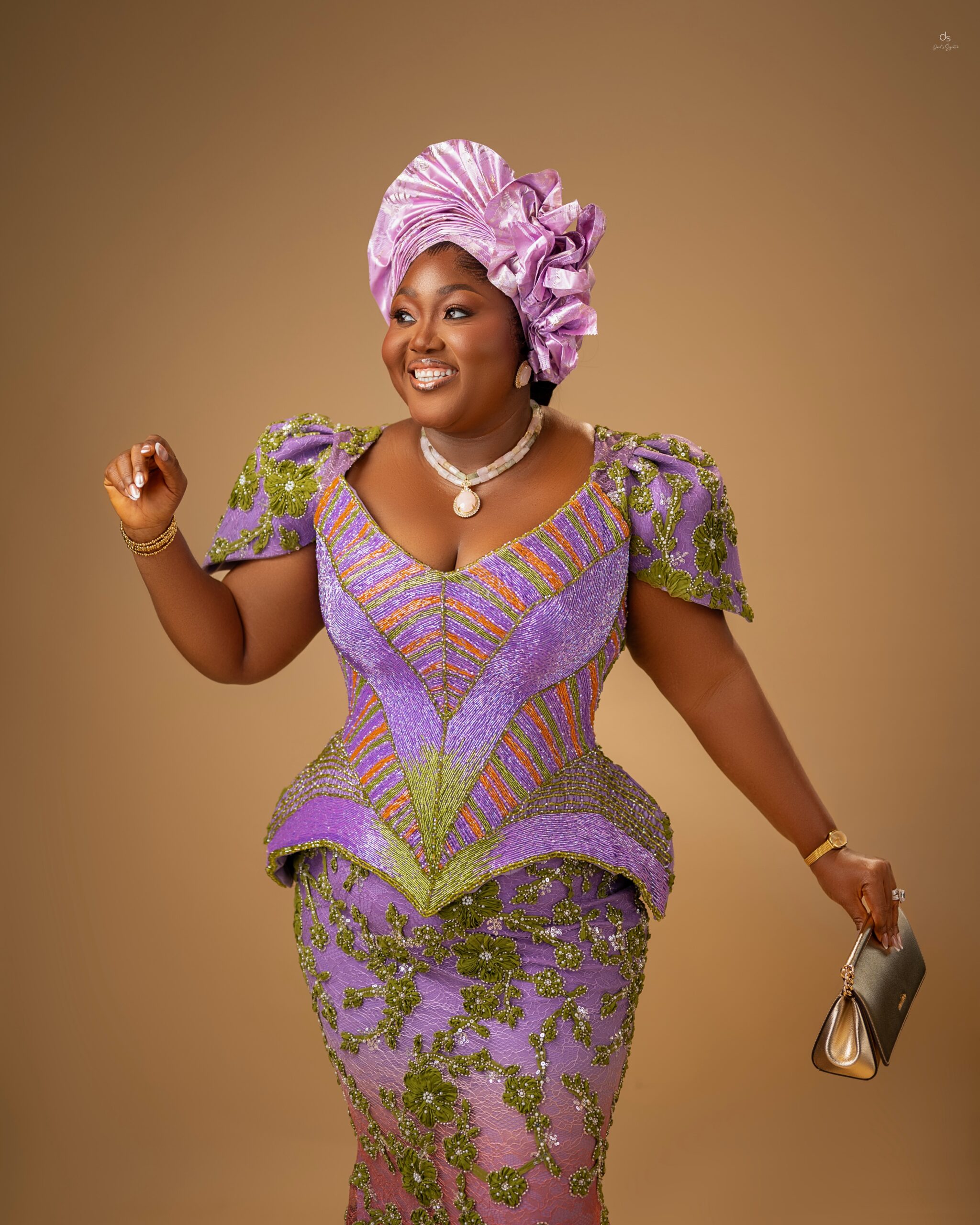 Blessing Nze smiling while wearing an ombré purple lace dress with green floral embroidery, a corset waist, and a matching lilac fan-pleated Gele headtie.