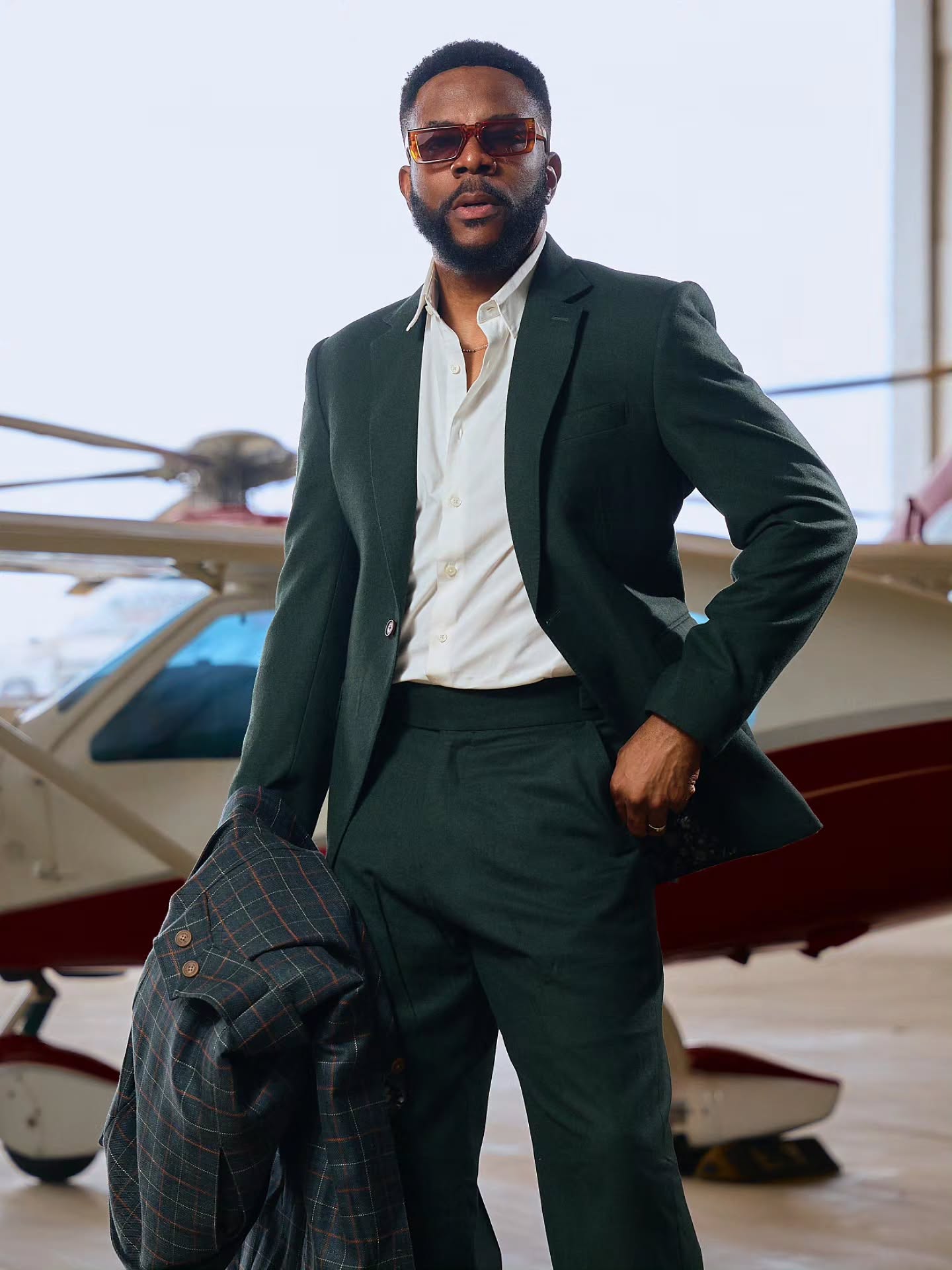 Ebuka Obi-Uchendu wearing a forest-green two-piece suit by Deji & Kola with high-waisted trousers and an unbuttoned white shirt, holding a plaid trench coat in an airfield hangar.