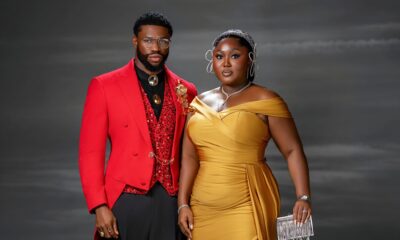 Blessing Jessica Obasi wearing a gold satin dress with a crystal Alice headband next to Stan Nze in a red tailcoat.