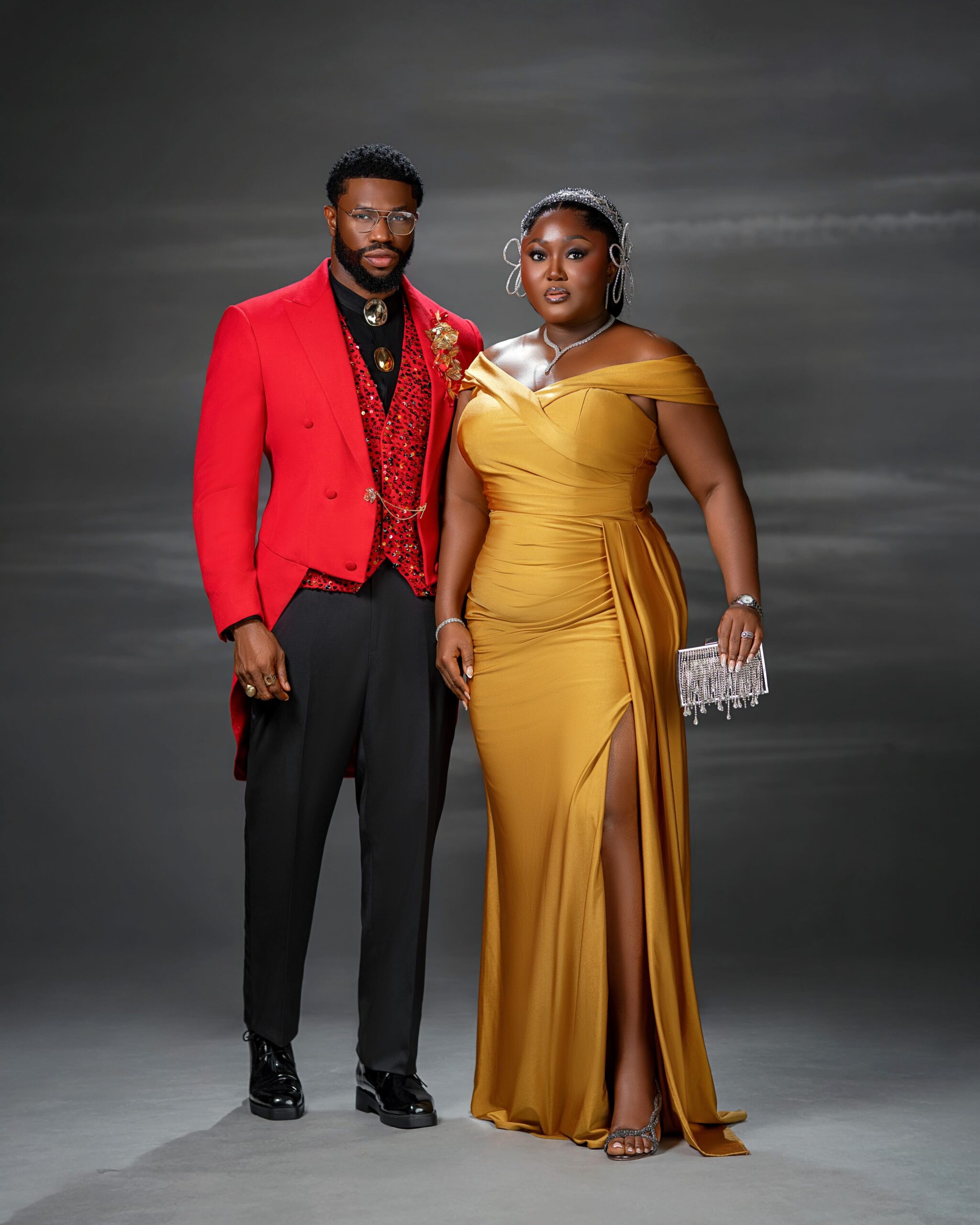 Blessing Jessica Obasi wearing a gold satin dress with a crystal Alice headband next to Stan Nze in a red tailcoat.