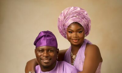 Olamide in a lilac corset halter gown hugging Kamo State wearing a lilac embroidered agbada from behind.