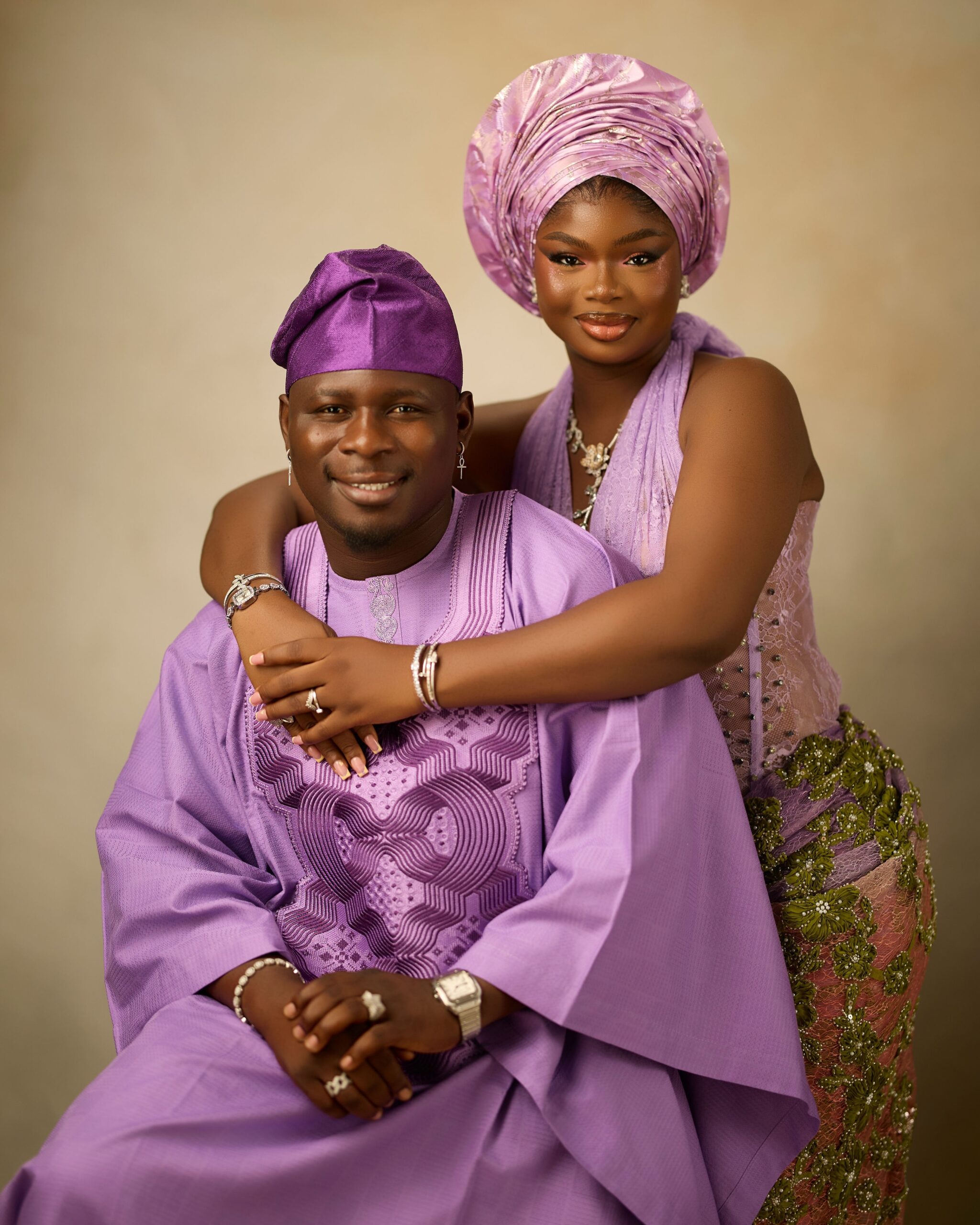 Olamide in a lilac corset halter gown hugging Kamo State wearing a lilac embroidered agbada from behind.