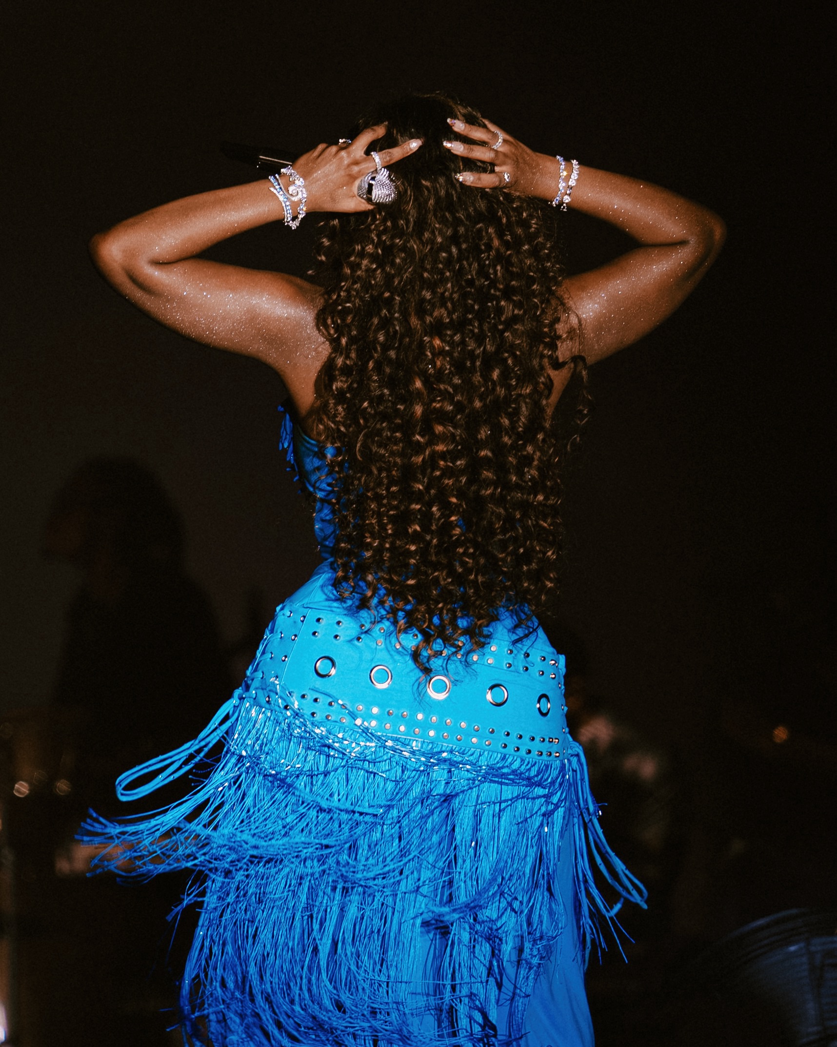 Back view of Tems with long dark brown curls wearing an electric blue dress with silver grommet waist details.