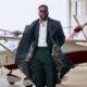 Ebuka Obi-Uchendu opening a charcoal windowpane Deji & Kola trench coat to reveal a floral silk interior lining over a green suit.