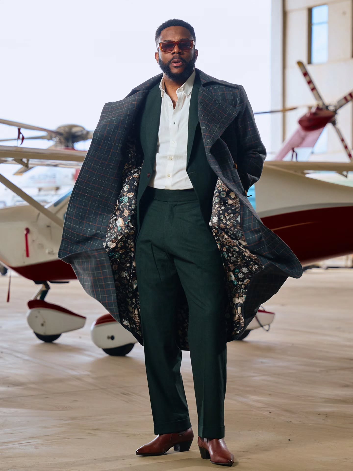 Ebuka Obi-Uchendu opening a charcoal windowpane Deji & Kola trench coat to reveal a floral silk interior lining over a green suit.