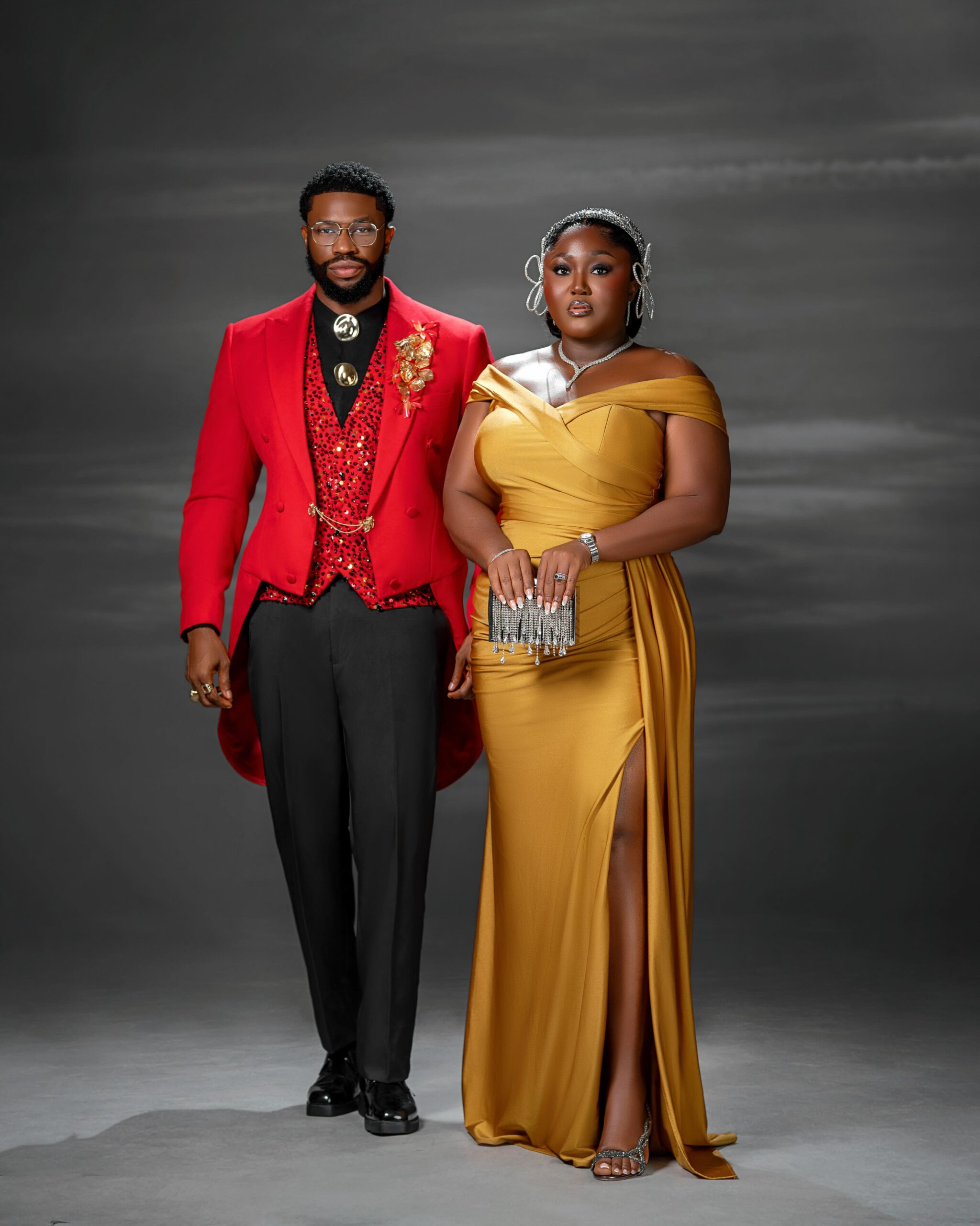 Stan Nze in a red double-breasted tailcoat posing alongside Blessing Jessica Obasi in a gold satin off-the-shoulder gown.