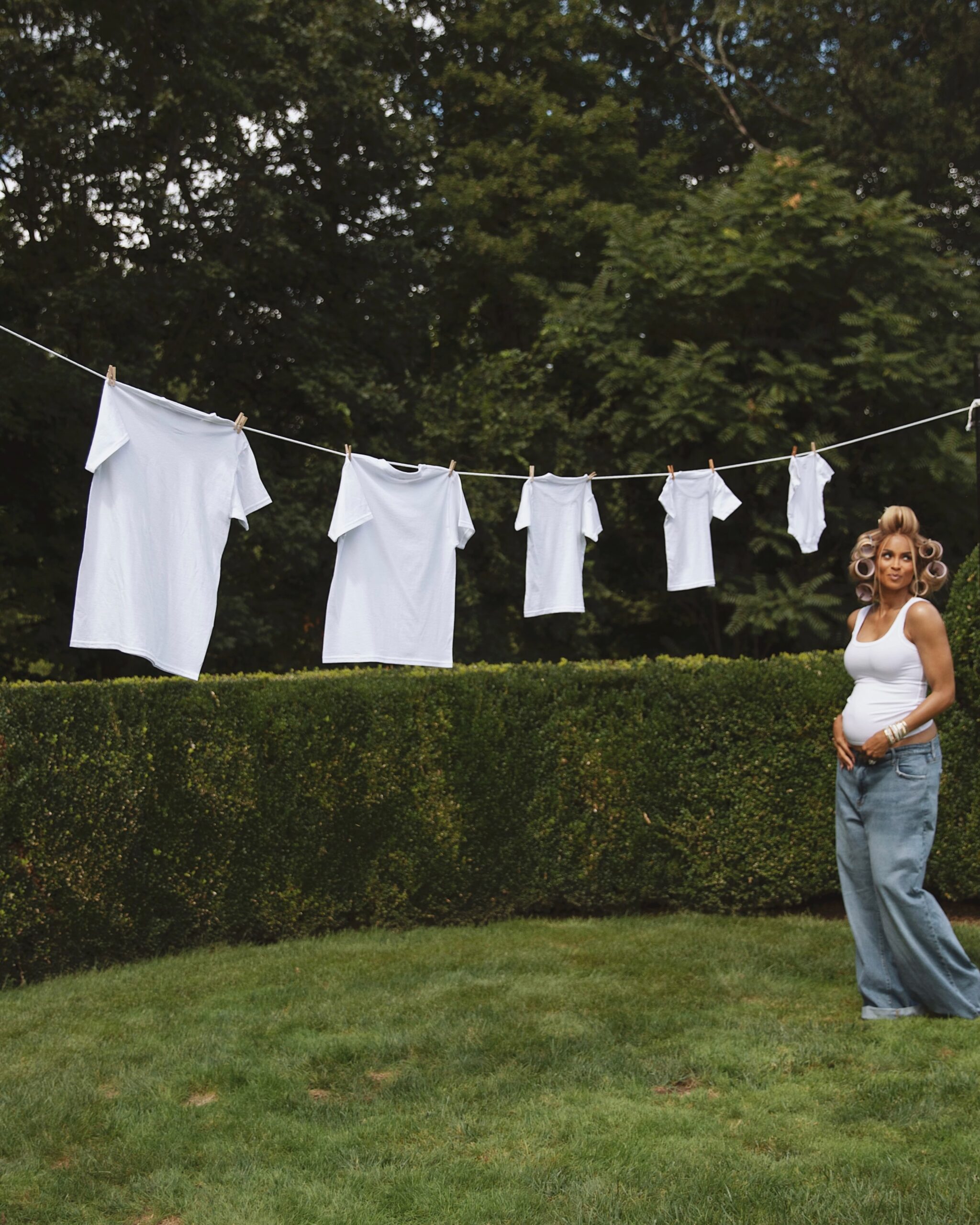 Ciara holding her baby bump next to a clothesline with five white shirts hanging in decreasing sizes.